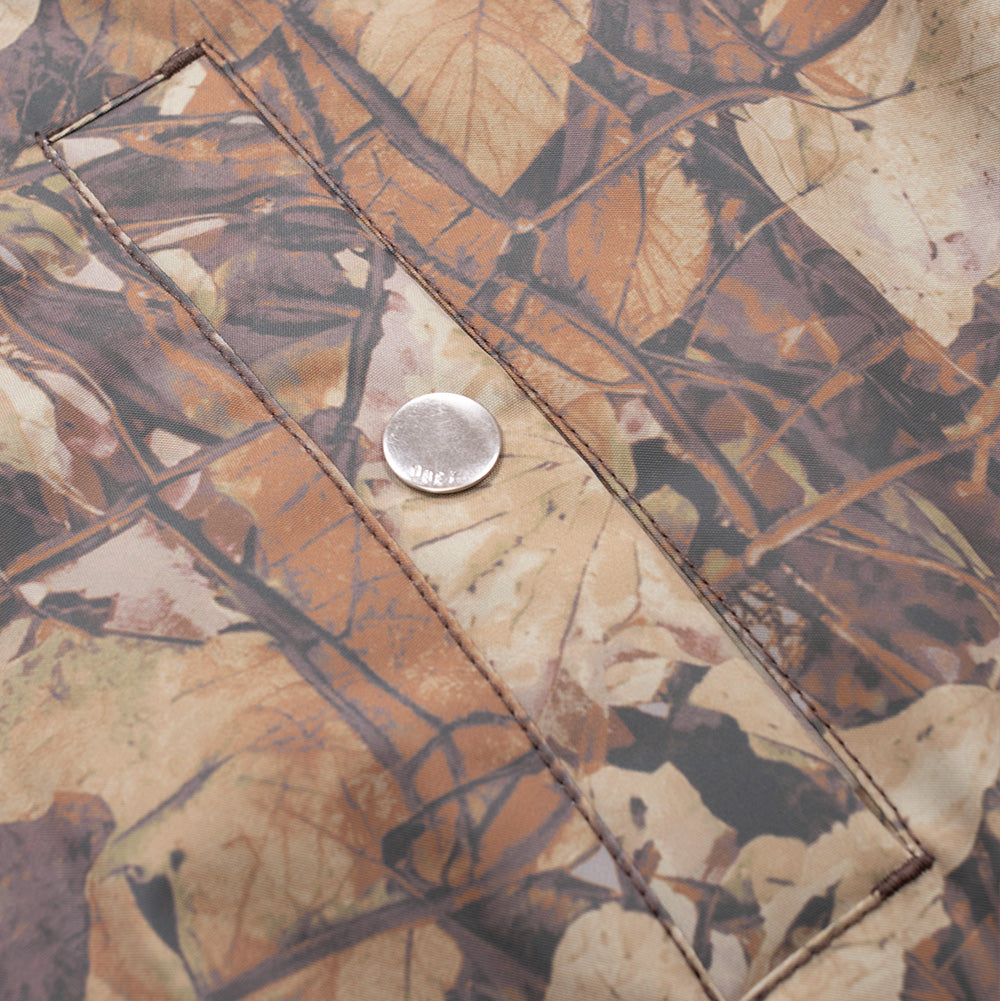 Macro perspective of the lower front pocket on the Obey N-2B Short Parka, showcasing the wide welt flap pocket construction with reinforced stitch lines and an OBEY-branded silver metallic snap fastener set on the padded camouflage shell.
