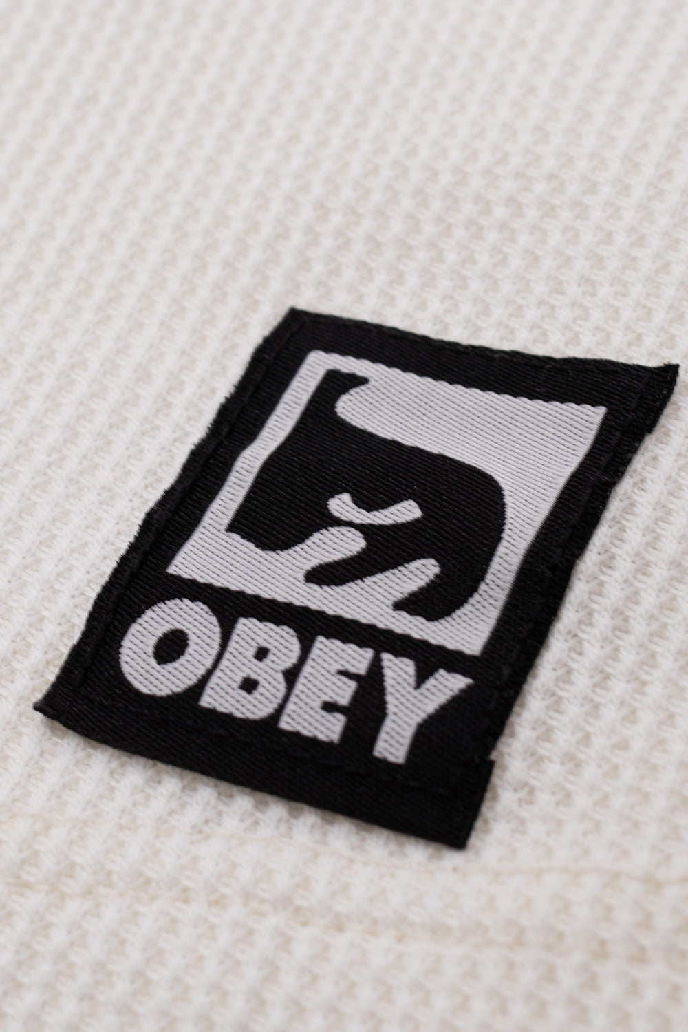 Macro close-up shot of the lower left hem on the Dusty Chalk Obey Post Thermal, showing the raised square grid weave of the coarse cream waffle-knit fabric and the sewn-on black and grey woven Obey patch featuring the iconic André the Giant face graphic and serif brand lettering.