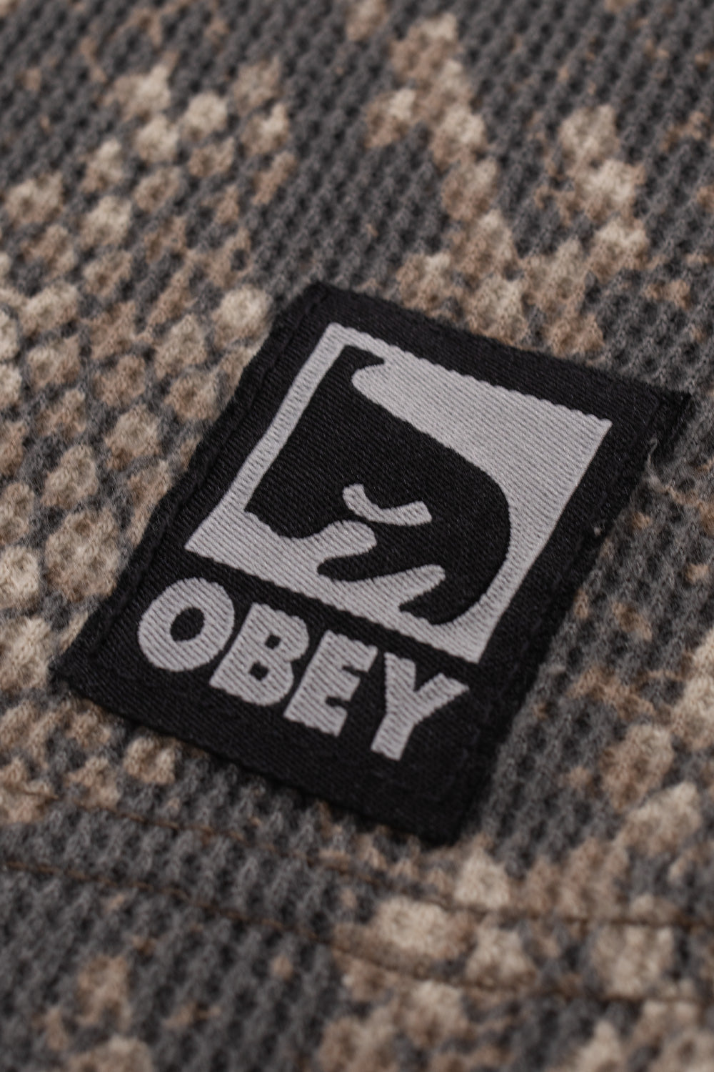 Macro close-up shot of the lower left hem on the Obey Post Thermal in Dusty Vintage Rattlesnake, highlighting the textured three-dimensional waffle knit grain with detailed python reptile scale printing and the sewn-on square black and grey Obey patch featuring the iconic André the Giant face graphic.