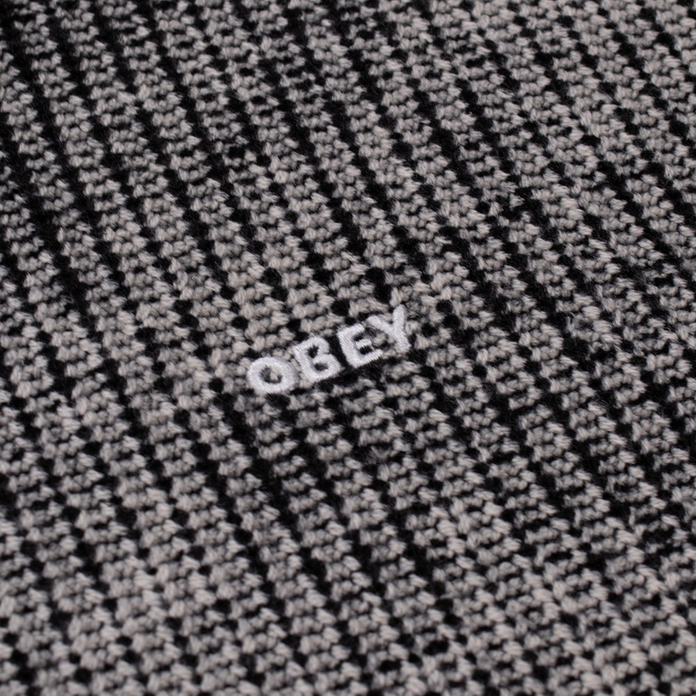 Macro studio close-up of the chest embroidery on the Obey Spray Effect Sweater, detailing the dense white satin-stitched "OBEY" block capital lettering centered on the textured two-tone marled black and grey ribbed knit yarn.
