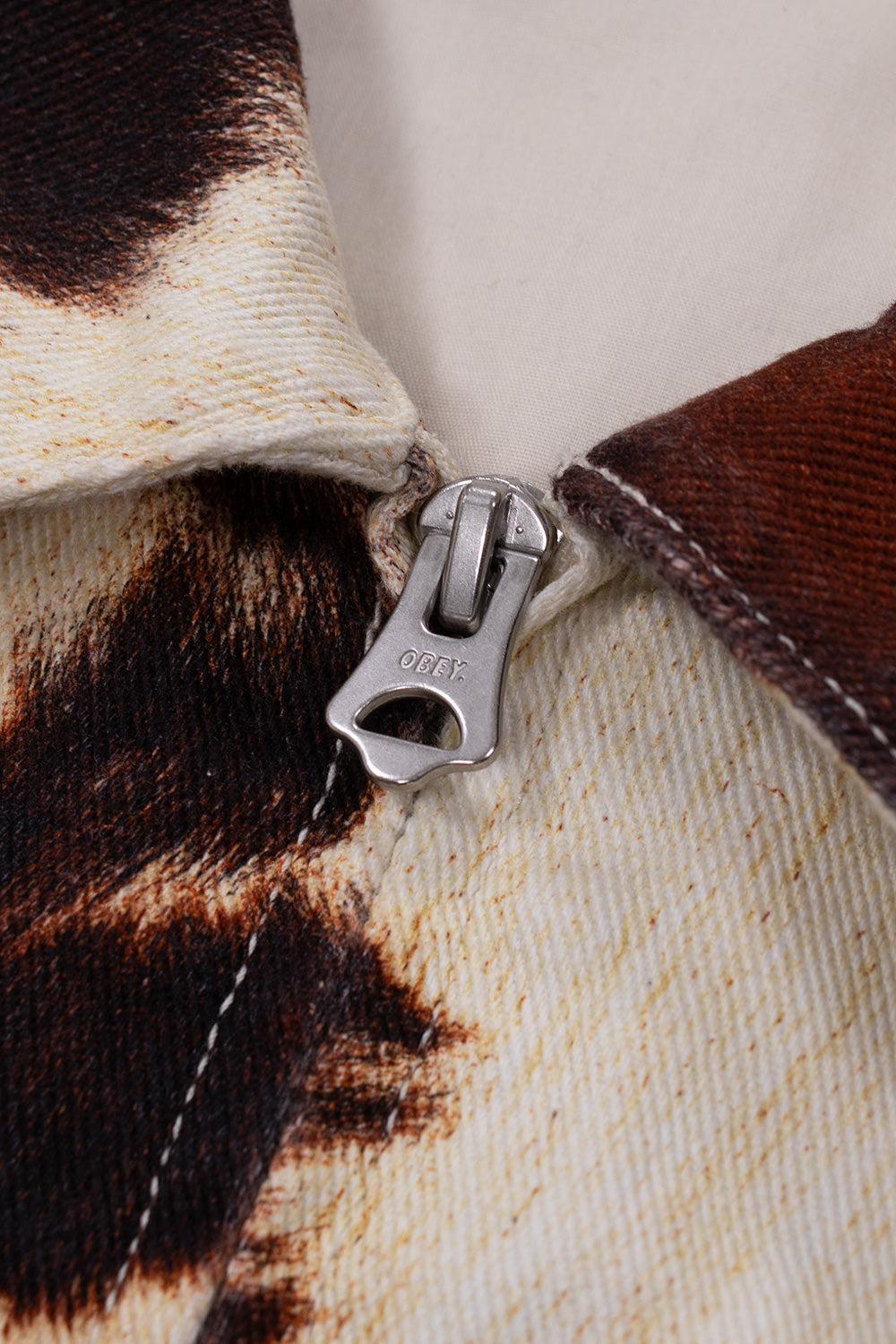 Macro close-up shot of the antiqued silver zipper pull engraved with the Obey brand text logo, positioned on the front collar seam of the 13.5oz printed cotton denim Workshop Jacket.