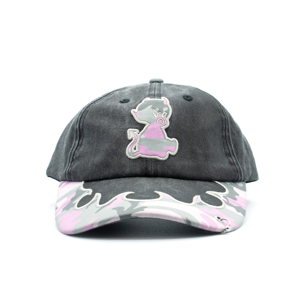 Front studio view of the Token Bae Cap, showcasing a washed faded black cotton six-panel dad hat crown, an embroidered center patch of a seated baby devil filled with pink camouflage pattern, and a curved curved visor detailed with pink-and-grey camo print, white flame border embroidery, and a silver metallic piercing ring on the brim.