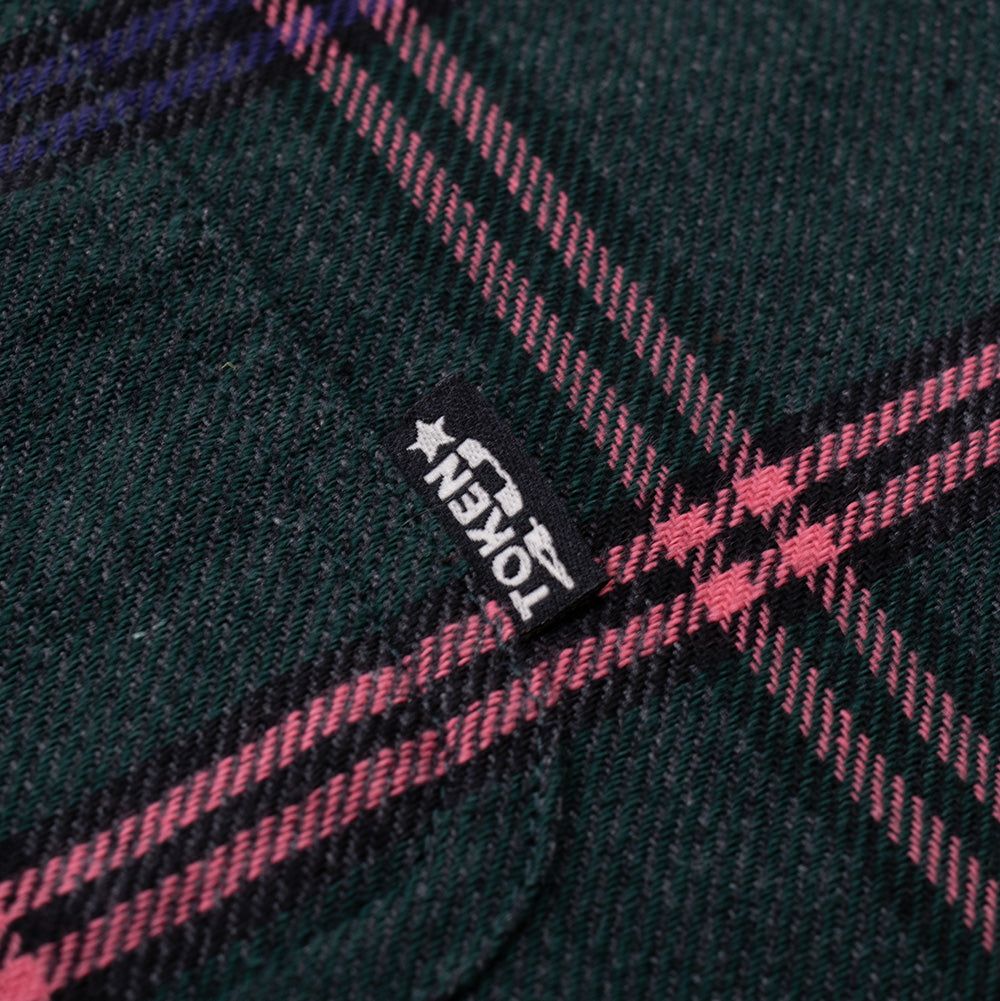 Extreme macro close-up of the woven brand flag label on the Token Hardware Plaid Shirt, showcasing the embroidered white "TOKEN" block text with star motif bordered by diagonal pink twill weave lines against the deep midnight green fabric.