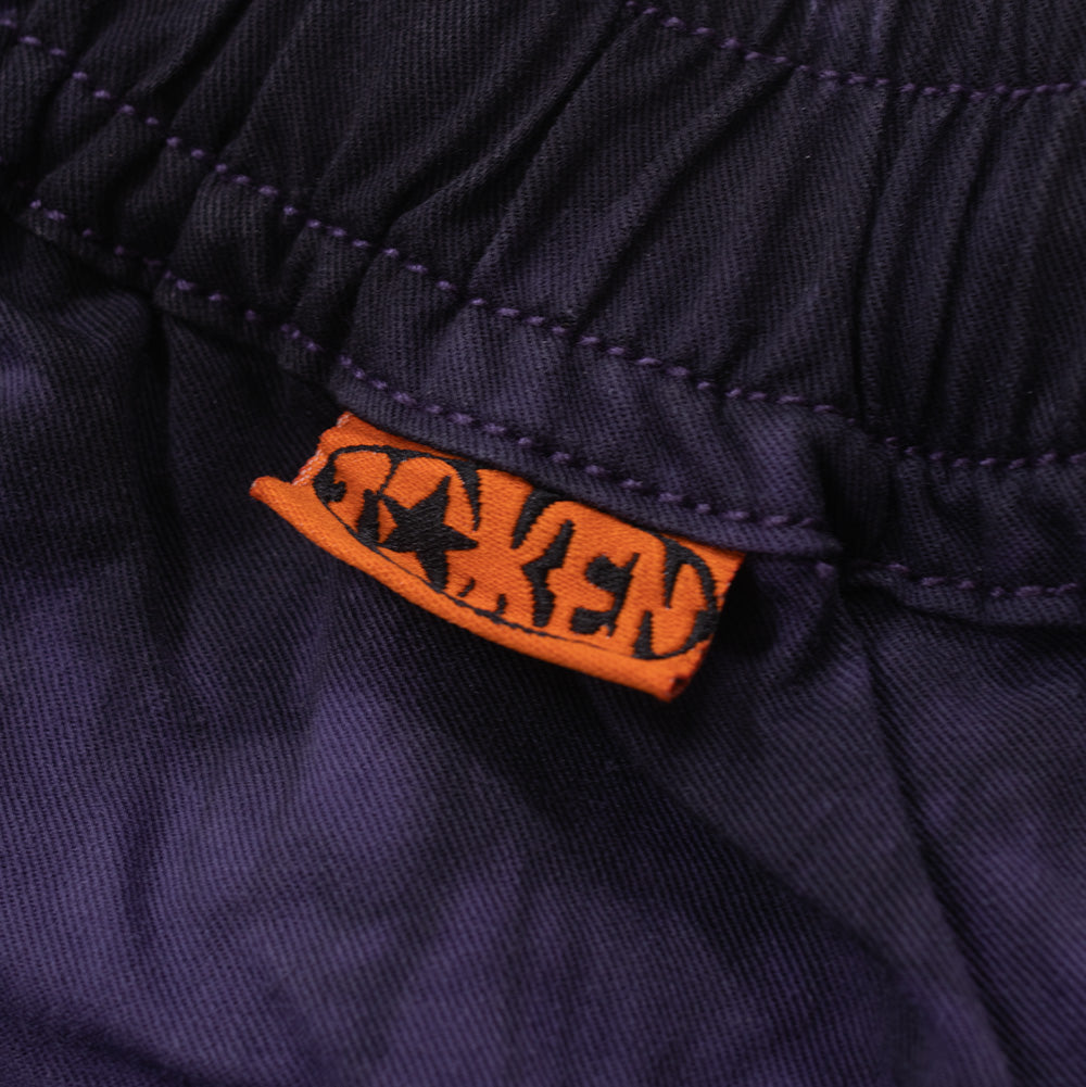 Extreme macro view of the waistband seam on the Token Sundown Pants, focusing on the vibrant neon orange mini woven brand tag featuring the black printed "TOKEN" wordmark with star detailing on the gathered purple fabric.