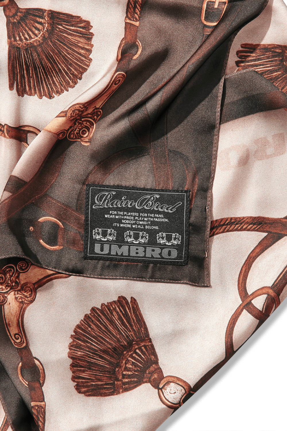 Macro studio close-up of the woven black label sewn along the folded hem of the Umbro Belt Masked Foulard, featuring white embroidered script reading "Rare Breed" above brand manifesto text and the Umbro wordmark flanked by double crest lions, set against smooth brown and cream printed polyester twill