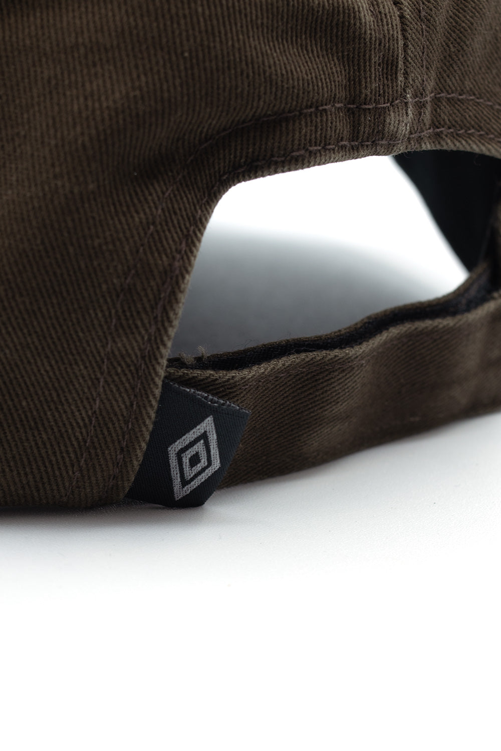 Detailed close-up shot of the rear adjustment strap on the Umbro Censored Cap, focusing on the folded woven black flag label displaying the silver Umbro double diamond logo attached to the washed brown cotton Velcro fastening band