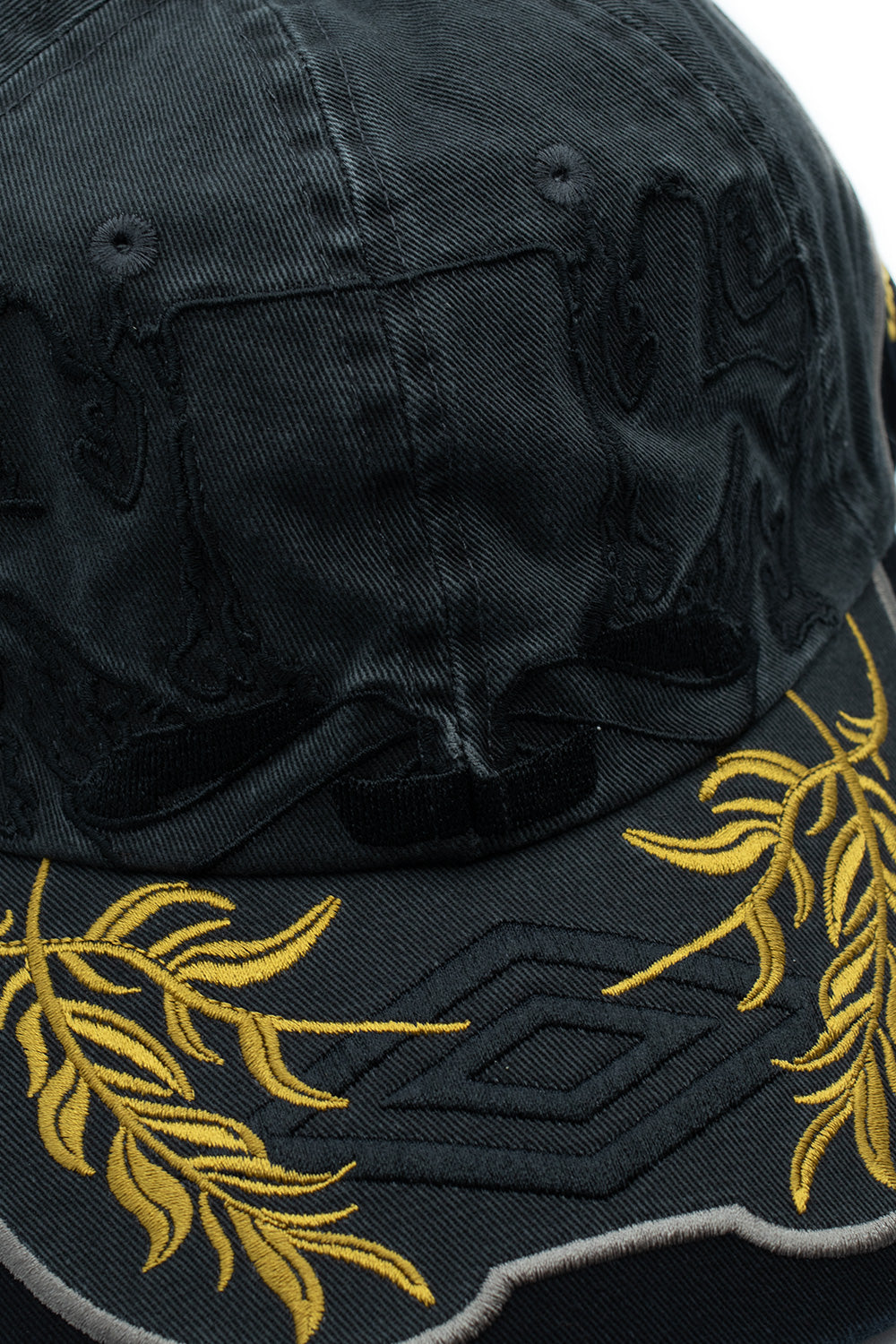 Macro close-up shot of the visor and lower crown seam on the Umbro Laurel Cap, detailing the dense golden-yellow satin stitching of the ornate laurel leaves, central black embroidered Umbro diamond, and tonal embroidered rampant lions on the washed dark olive-green fabric