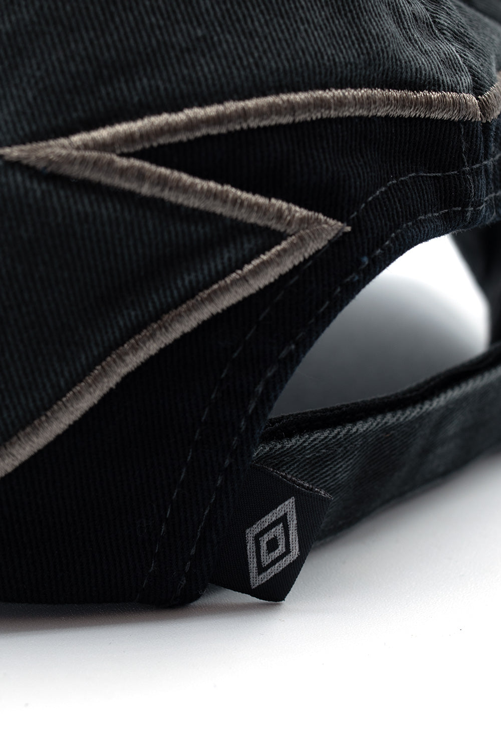 Detailed studio close-up of the rear closure on the Umbro Laurel Cap, focusing on the folded black woven label featuring a silver Umbro double diamond logo attached to the washed green cotton strap alongside dense taupe border embroidery