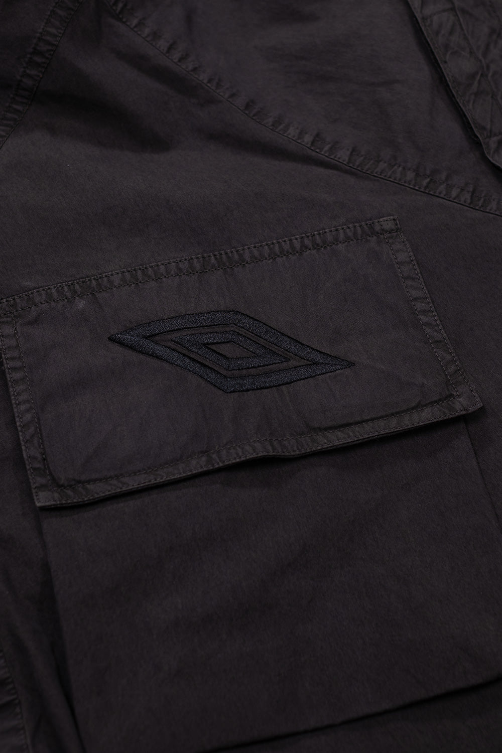 Close-up of Umbro Mid Field Jacket chest pocket with embroidered double diamond logo on dark olive garment-dyed fabric