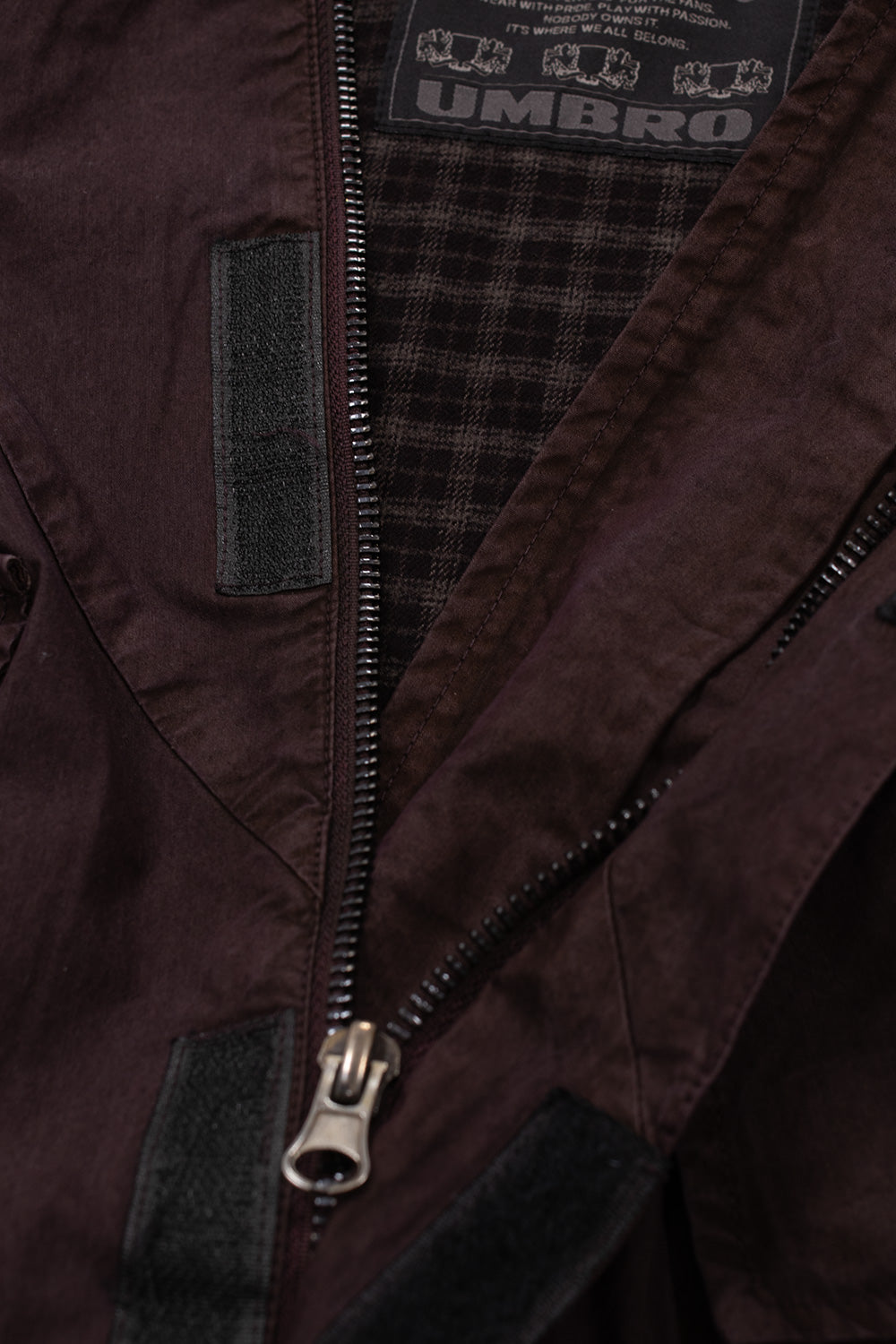 Detailed shot of the partially unzipped neckline on the Umbro Midfield Jacket, revealing the internal dark tartan plaid check fabric lining, woven Umbro heritage brand label at the inner back neck, and the robust metal zipper hardware