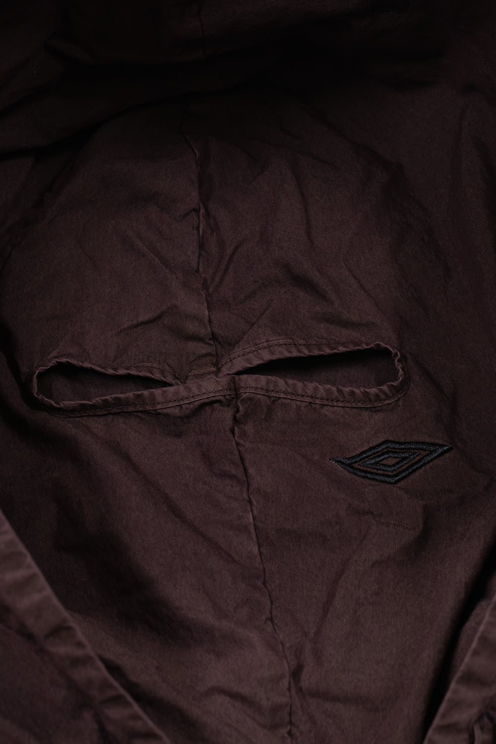 Detailed studio close-up of the hood on the Umbro Sunday Masked Jacket, highlighting the integrated matching plum-colored balaclava face panel with finished eye slits and a tonal embroidered Umbro double diamond logo on the cheek