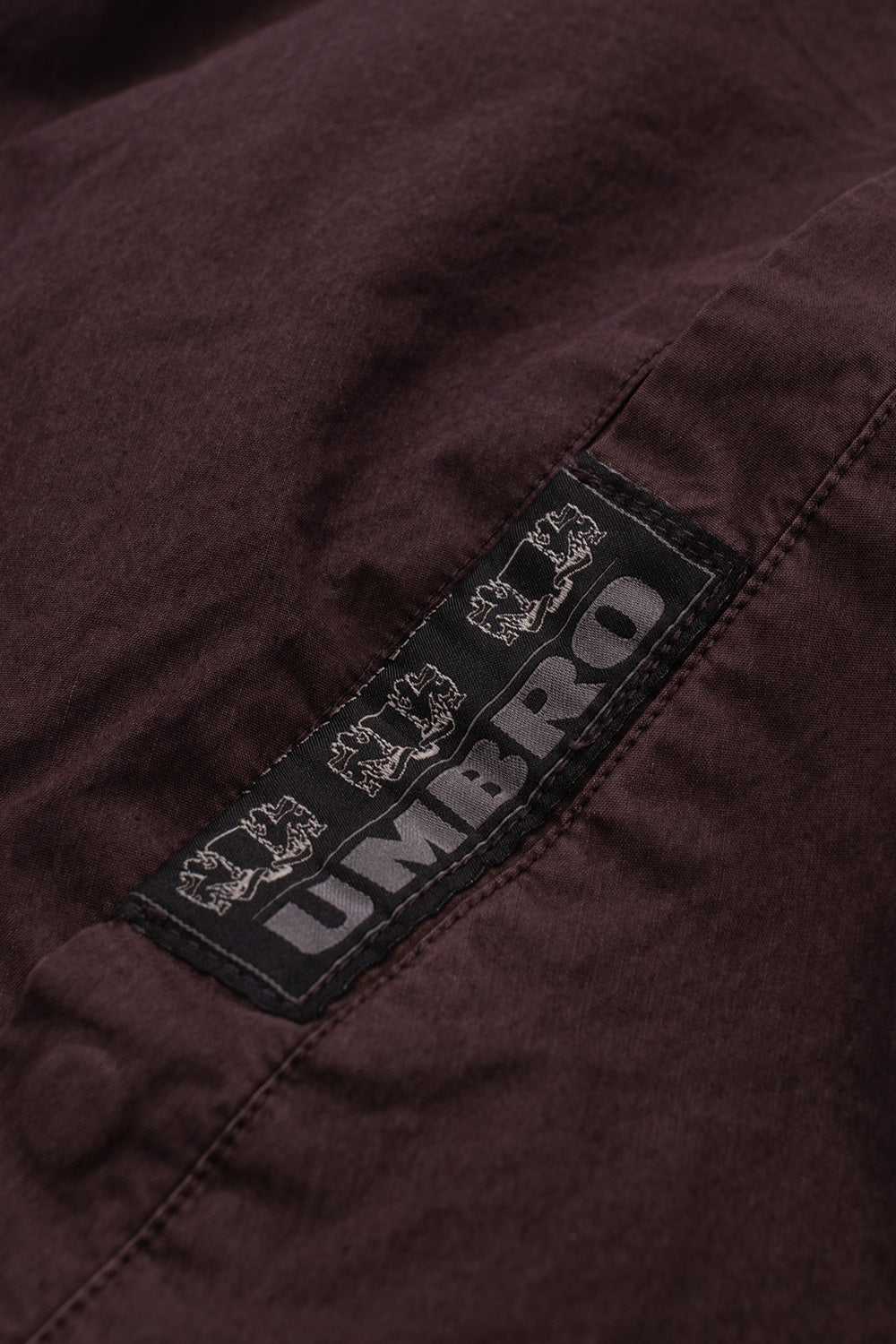 Macro close-up view of the black woven heritage label on the right pocket welt of the Umbro Sunday Masked Jacket, featuring grey printed "UMBRO" block lettering flanked by heraldic crest lions on the washed purple textile