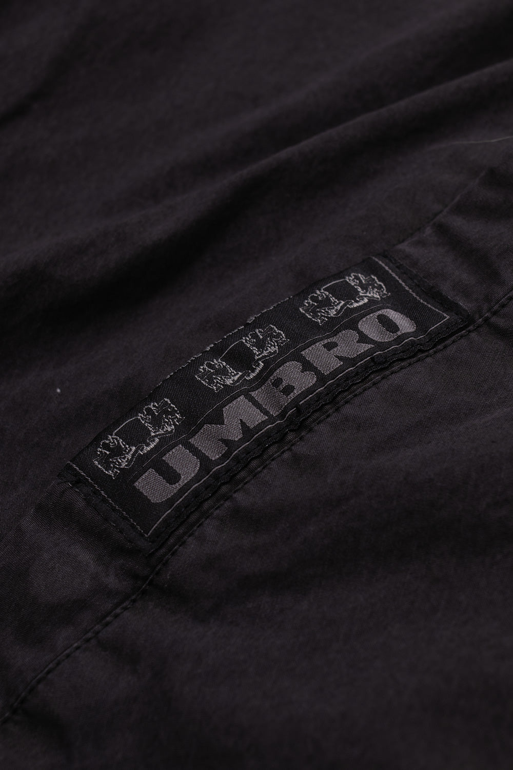 Macro close-up view of the black woven heritage label stitched onto the right pocket welt of the Umbro Sunday Masked Jacket, featuring grey "UMBRO" block lettering flanked by heraldic crest lions on the washed black cotton fabric
