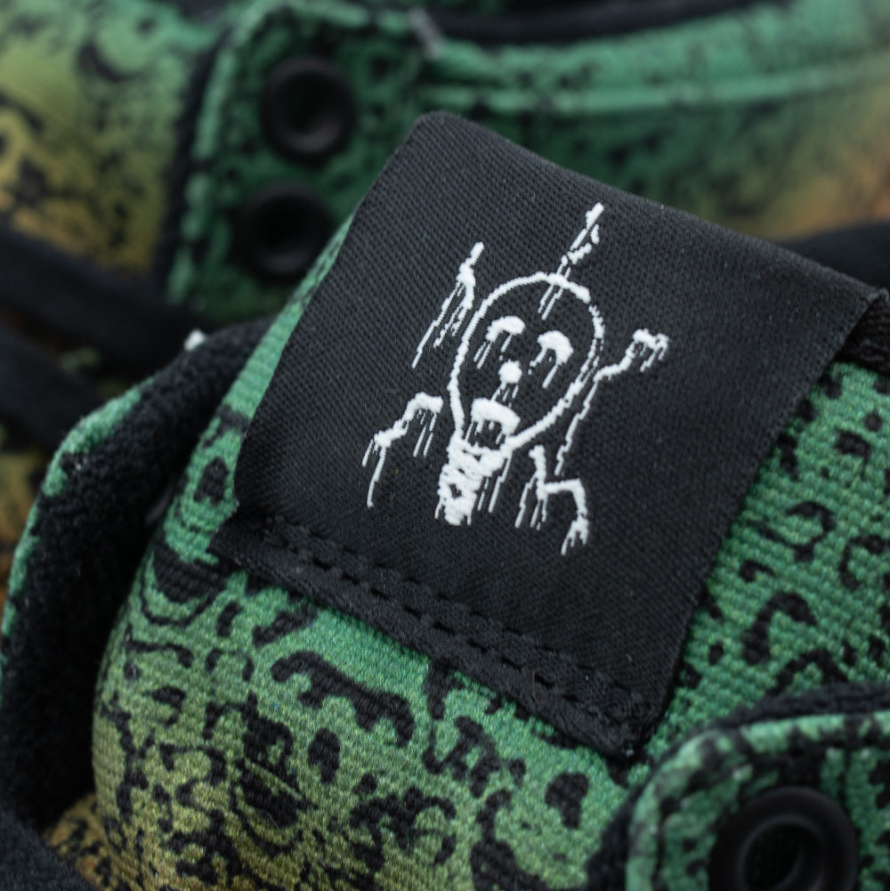 Macro studio close-up of the tongue label on the Vans OTW x Chito Half Cab 33, detailing the woven black fabric tag featuring Chito's iconic white dripping character sketch, set against the green skull-printed canvas tongue and framed by black eyelets and laces.