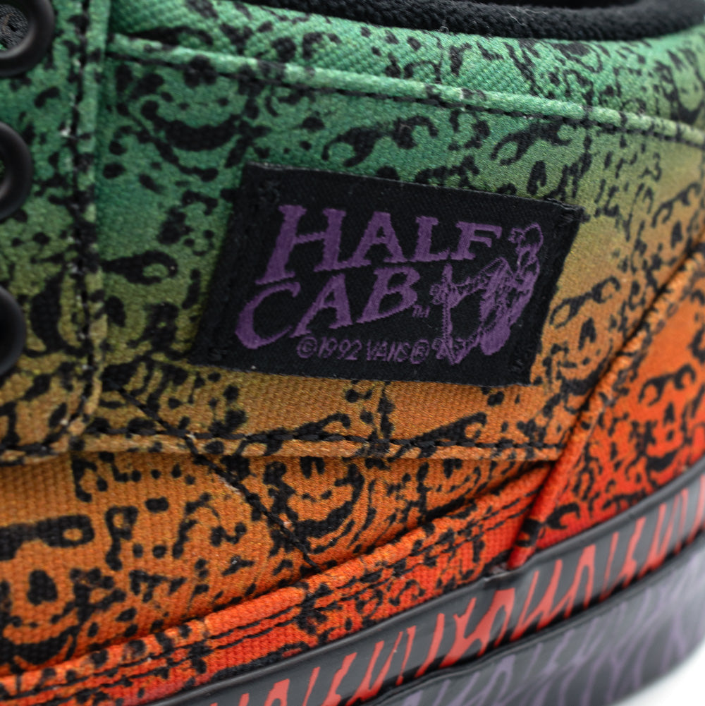 Detailed macro perspective of the lateral ankle on the Vans OTW x Chito Half Cab 33, focusing on the rectangular woven black label embroidered with purple "HALF CAB" typography and Steve Caballero's skateboarding silhouette, set over the green-to-orange gradient skull canvas and red tribal foxing tape.