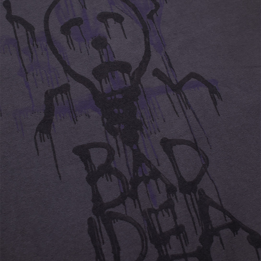 Detailed macro perspective of the front artwork on the Vans OTW x Chito T-Shirt, capturing the spray-paint texture of the black dripping lightbulb creature graphic set over faint purple crosshair overspray and dripping "BAD IDEA" lettering on faded black cotton fabric.