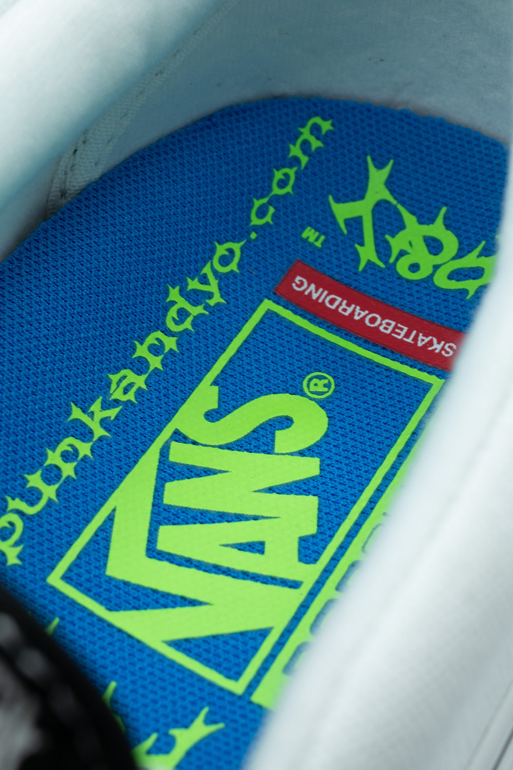 Detailed interior macro shot of the Vans X Punk And Yo Skate Slip-On shoe, showing the electric blue PopCush footbed insole printed with a large neon lime green Vans Skateboarding logo, gothic green "punkandyo.com" URL lettering, "P&Y TM" branding, and the padded white leather inner collar lining