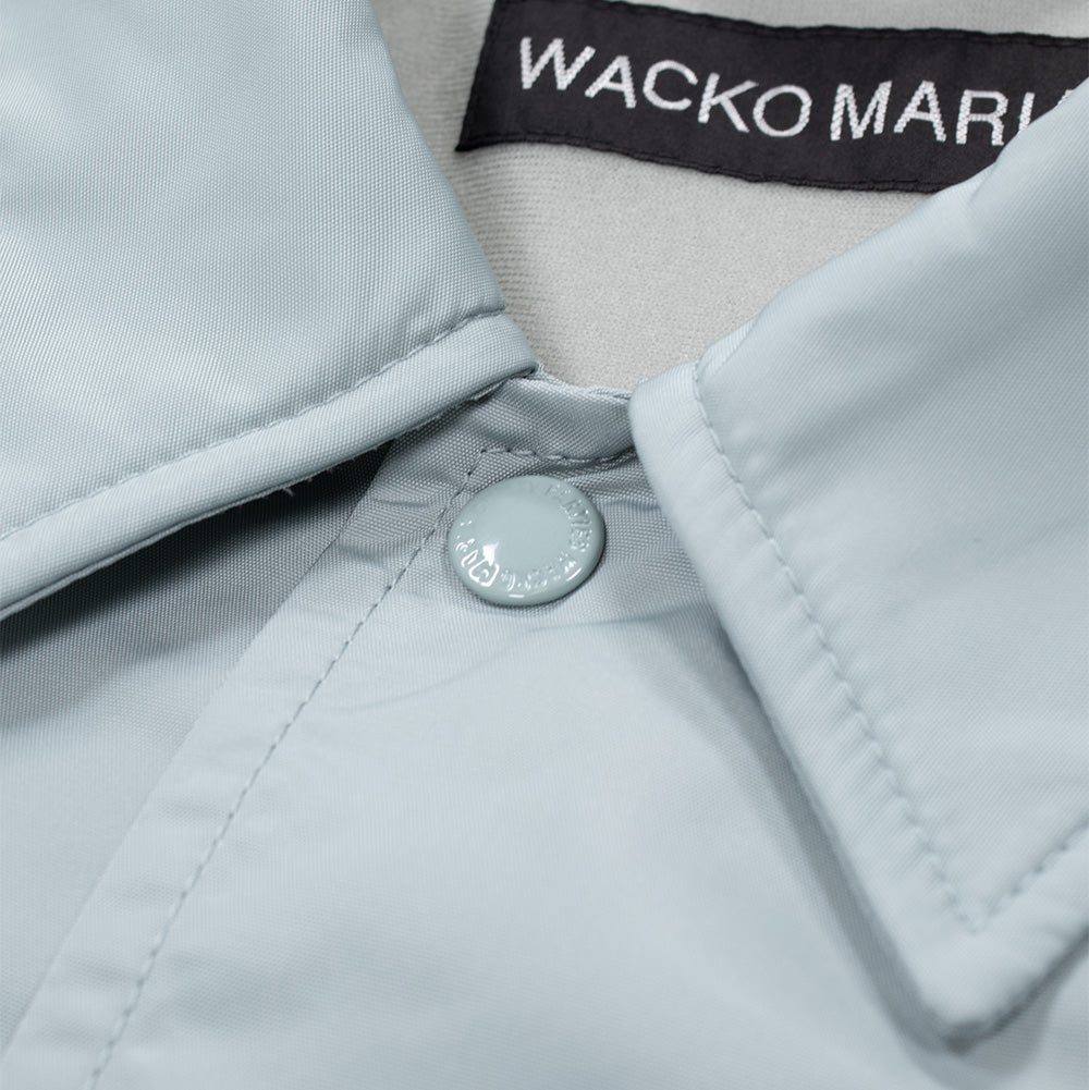 Macro studio close-up of the upper neckline and collar on the grey Wacko Maria Coach Jacket, displaying the smooth nylon-look shell textile, tonal enamelled metal snap button engraved with "GUILTY PARTIES WACKO MARIA", and the woven black brand tag along the inner collar seam.