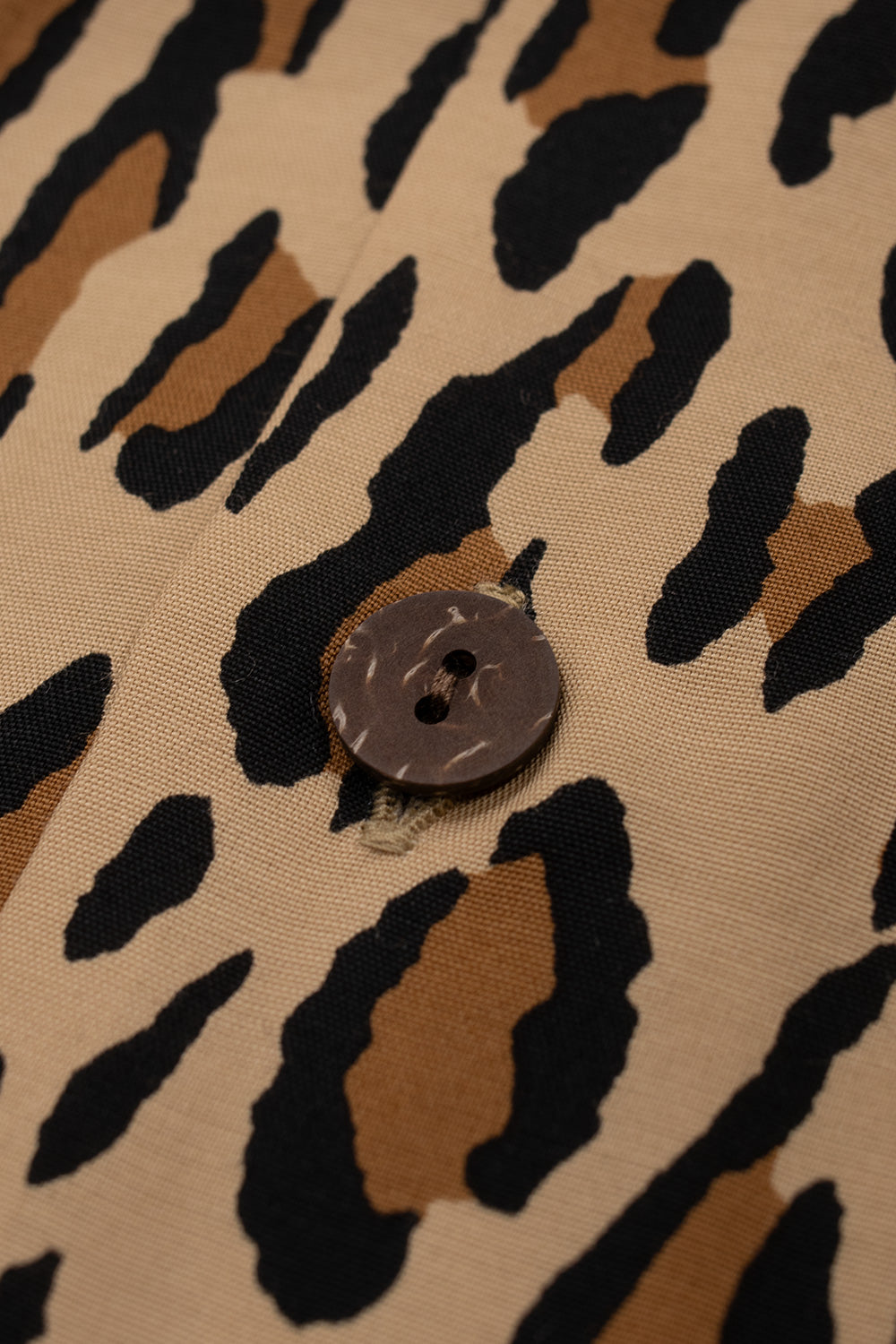 Macro view of a natural round wooden button stitched onto the button placket of the brown Wacko Maria rayon Hawaiian shirt.