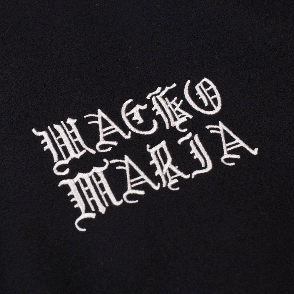Detailed macro perspective of the chest embroidery on the Wacko Maria Leather Varsity Jacket, highlighting the dense white satin-stitch thread forming the two-tier gothic Old English "WACKO MARIA" brand logo against smooth black wool fabric.