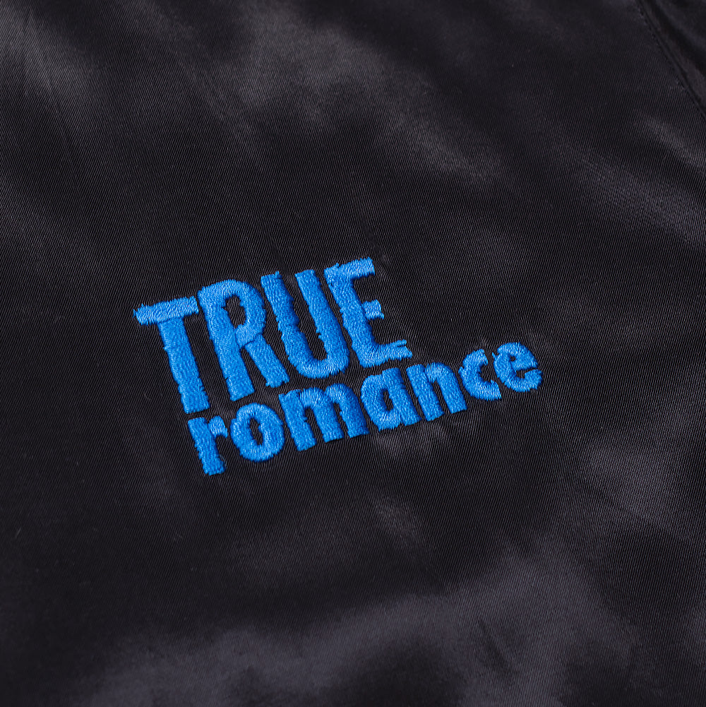 Detailed macro perspective of the chest embroidery on the black satin Wacko Maria x True Romance Ska Jacket, showing the textured turquoise blue satin-stitch thread forming the two-tiered "TRUE ROMANCE" movie title font.