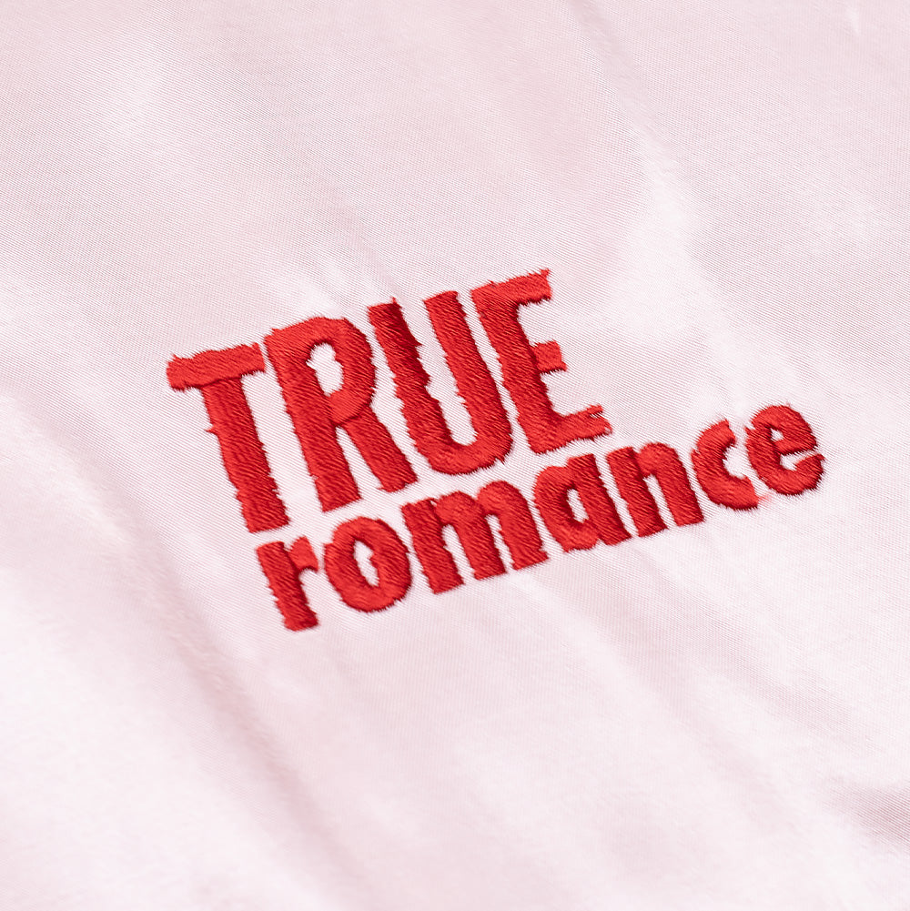 Extreme macro close-up of the front logo on the pink satin side of the Wacko Maria x True Romance Ska Jacket, highlighting the dense, vibrant red embroidered thread creating the stacked "TRUE ROMANCE" script against shimmering blush pink textile.