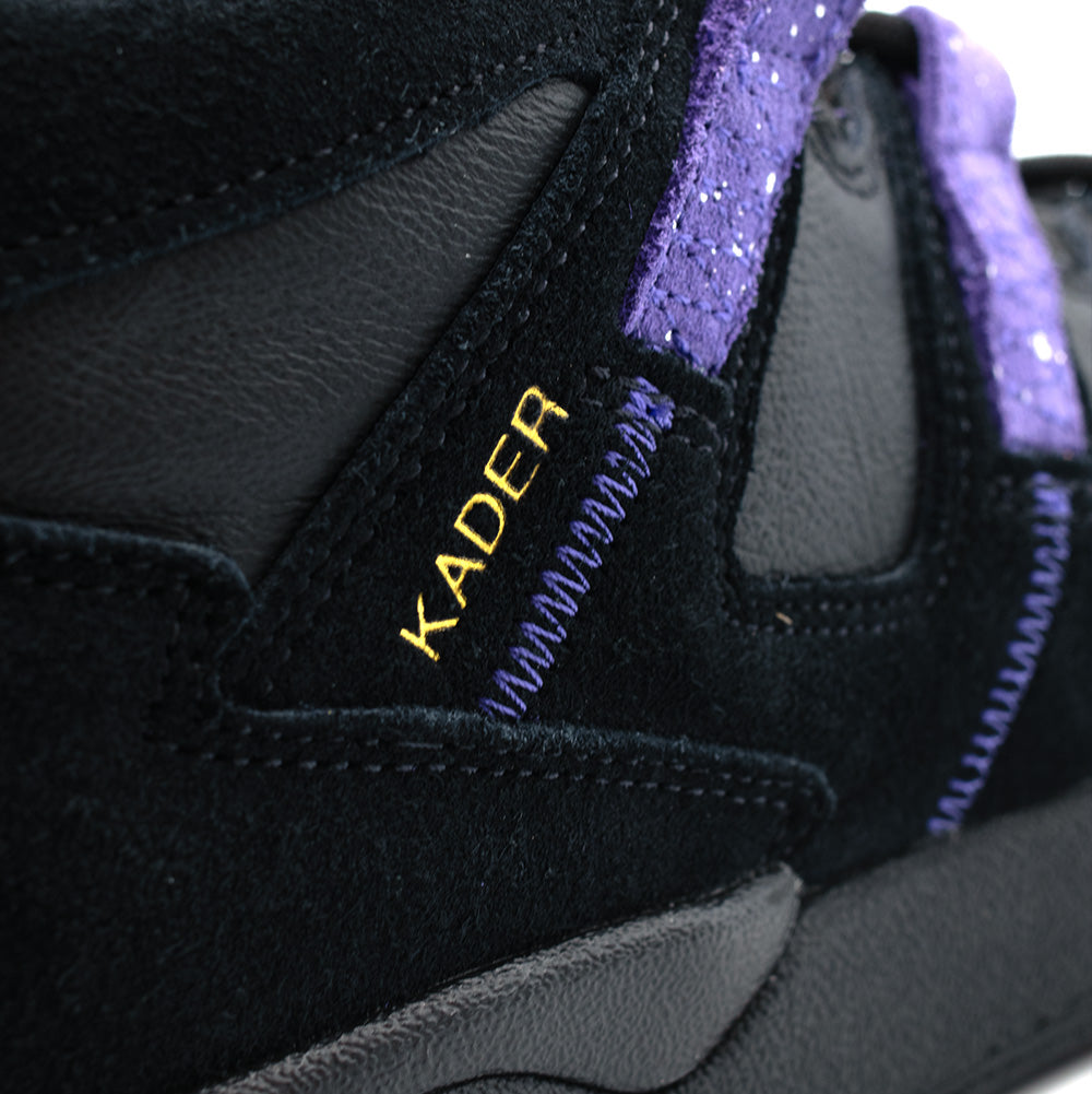 Macro perspective of the lateral ankle panel on the adidas Kader, showcasing the metallic gold foil "KADER" signature logo printed onto premium black suede, detailed with zig-zag purple contrast reinforcement stitching and the upper black cupsole edge.