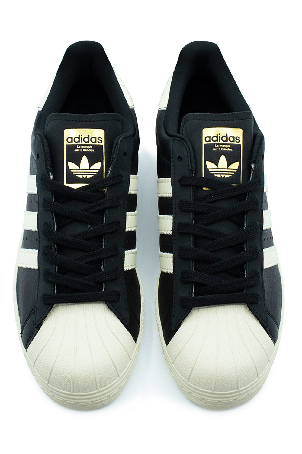 Top-down view of a pair of Adidas Superstar 80 ADV skate shoes, highlighting the cream white rubber shell toe box, black flat laces resting over subtle black nubuck eyestay overlays, and black leather tongues with gold-embossed Adidas Trefoil logo patches.