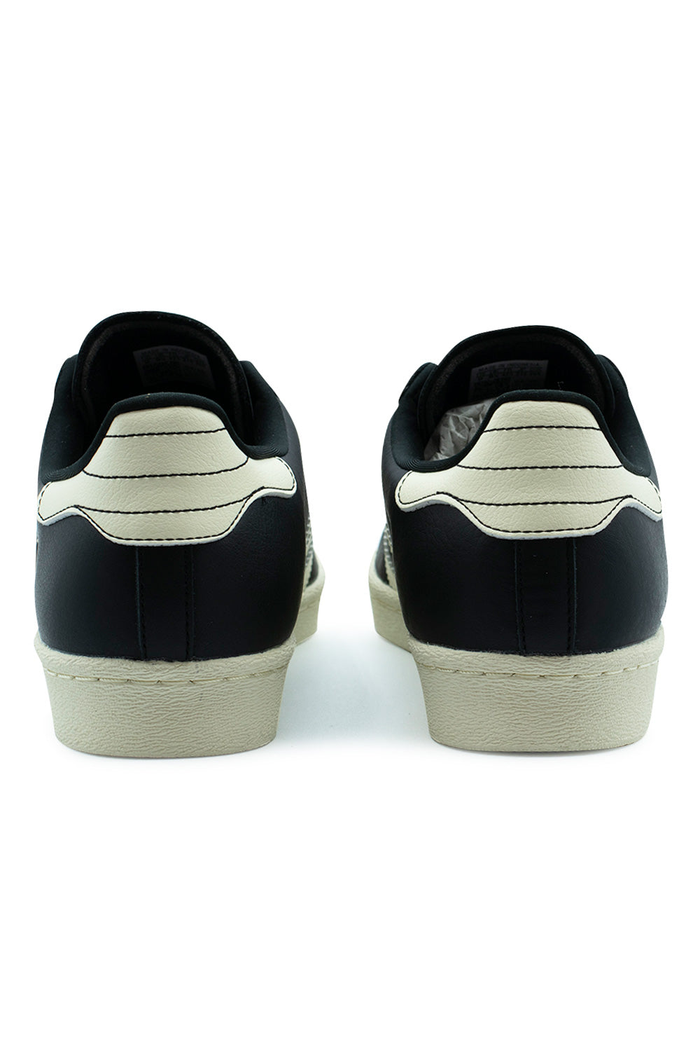 Rear view of the heels of the Adidas Superstar 80 ADV shoes, showcasing the black full-grain leather heel counters topped with cream white leather heel tabs featuring triple horizontal contrast stitching, resting on a textured cream cupsole.