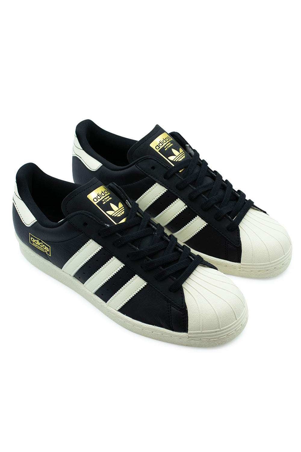 Three-quarter perspective studio shot of the Adidas Skateboarding Superstar 80 ADV shoes, demonstrating the classic 70s low-top silhouette, durable rubber toe bumper, black leather quarter panels, and gold-stamped side logo box.