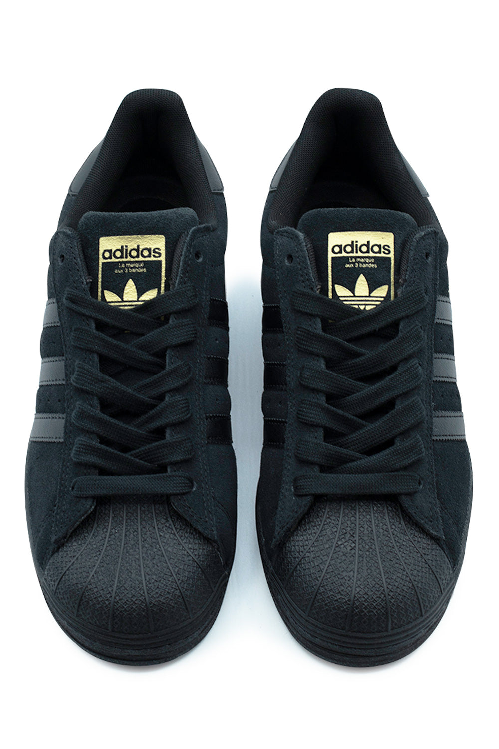 Top-down perspective of a pair of black Adidas Superstar ADV skate shoes, highlighting the durable black rubber shell toe boxes, flat black cotton laces, and black suede tongues with metallic gold foil Trefoil branding.