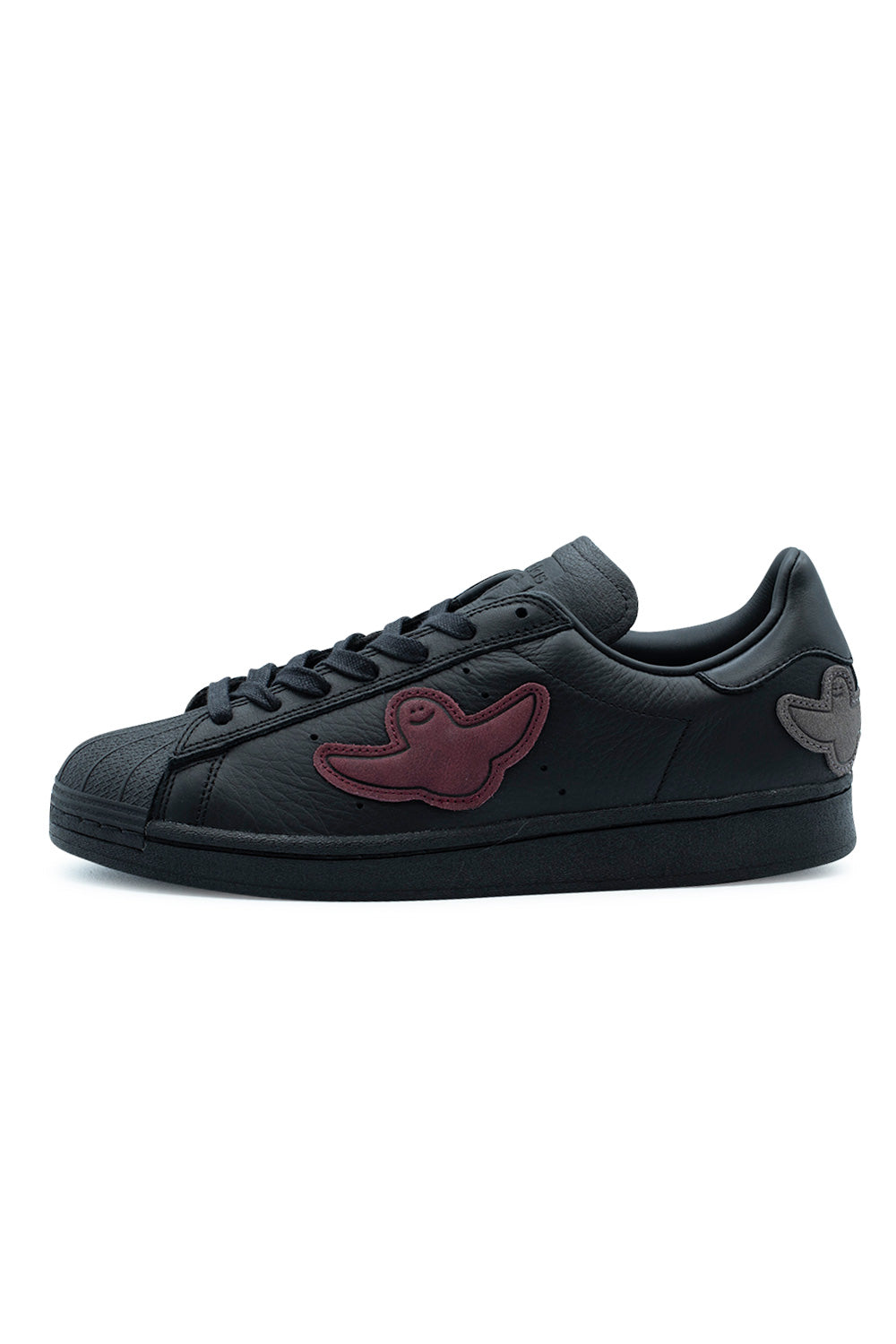 Lateral side profile of the Adidas Superstar ADV skate shoe by Mark Gonzales in Core Black, showcasing premium full-grain black leather uppers, stitched burgundy and grey suede Shmoo ghost patches, tonal perforated 3-Stripes ventilation holes, a textured black rubber shell toe, waxed laces, and a stitched all-black rubber cupsole.