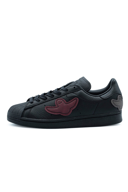 Lateral side profile of the Adidas Superstar ADV skate shoe by Mark Gonzales in Core Black, showcasing premium full-grain black leather uppers, stitched burgundy and grey suede Shmoo ghost patches, tonal perforated 3-Stripes ventilation holes, a textured black rubber shell toe, waxed laces, and a stitched all-black rubber cupsole.