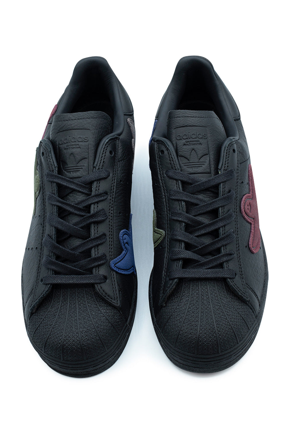 Top-down view of a pair of Adidas Superstar ADV Mark Gonzales shoes, displaying the iconic ridged rubber shell toe caps, flat black waxed laces, debossed Trefoil tongue badges, and asymmetric colorful suede Shmoo patches in olive green, burgundy, and cobalt blue across the eyestays and lateral sides.