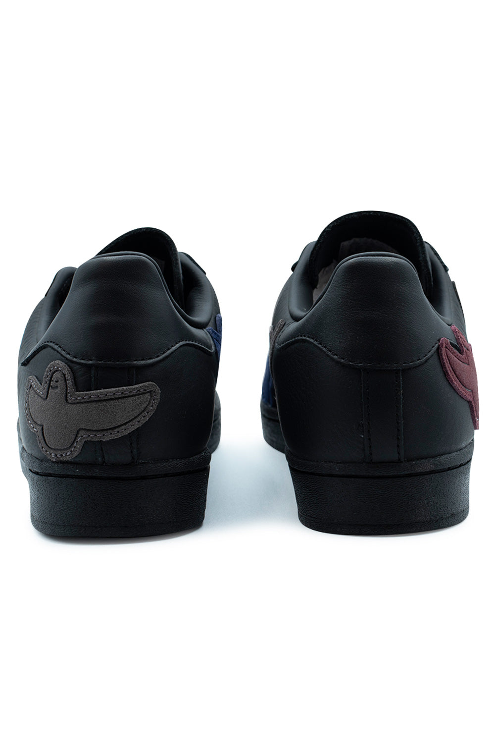 Rear perspective shot of the heels of the Adidas Superstar ADV Gonz sneakers, highlighting the smooth black leather heel tabs, tonal reinforcement stitching, solid black rubber midsoles, and an embroidered charcoal grey suede Shmoo character wrapping around the left heel counter.
