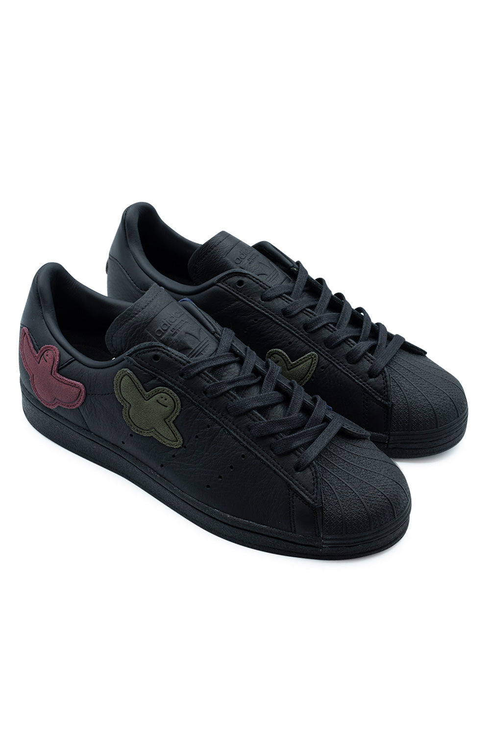 Elevated three-quarter studio view of the Adidas Superstar ADV Mark Gonzales in Core Black, highlighting the low-top skate silhouette, durable leather construction, protective rubber toe cap, and playful multi-colored suede Shmoo ghost appliques.