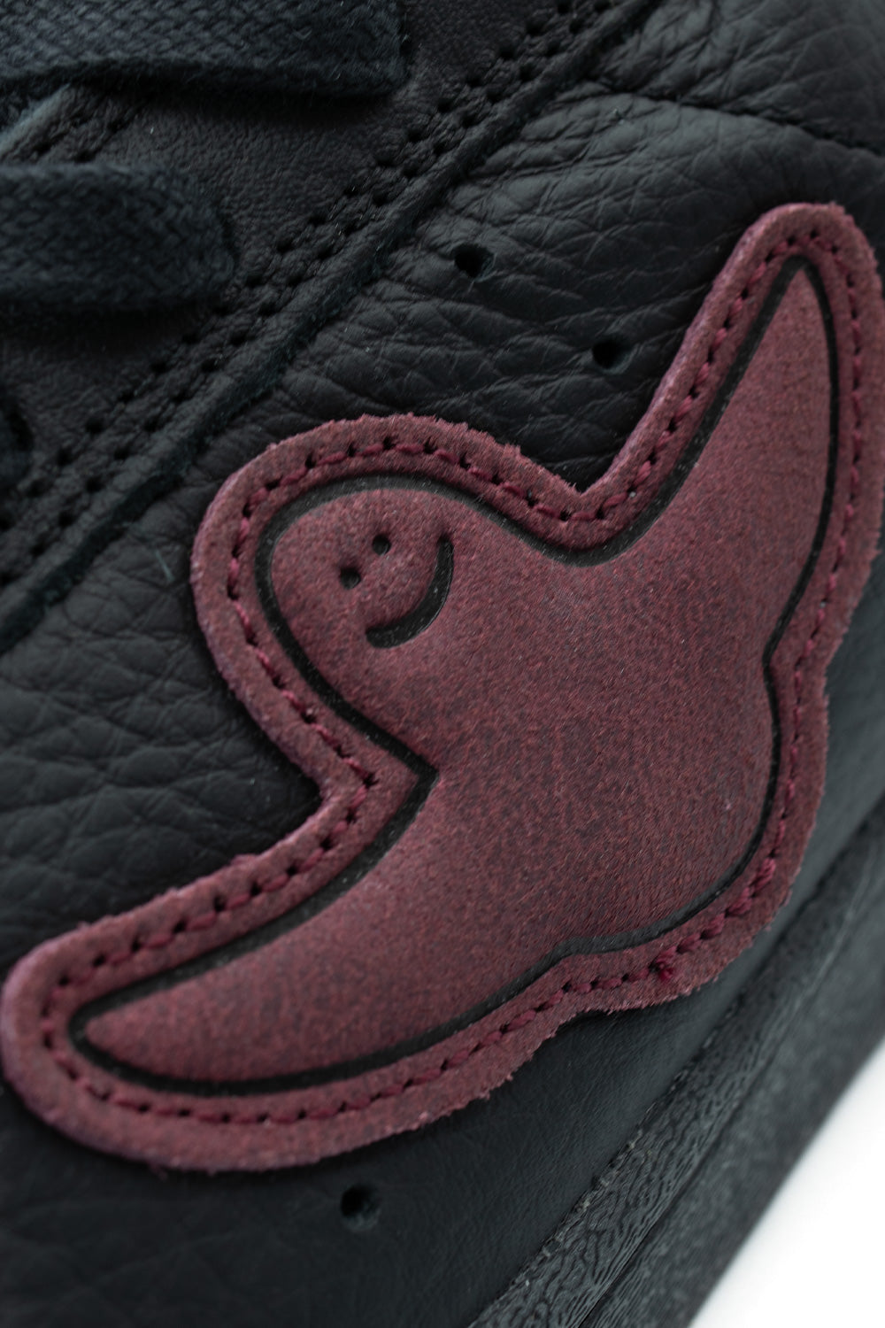 Macro close-up shot of the lateral side panel on the Adidas Superstar ADV Gonz, highlighting the textured grain of the black leather, subtle ventilation perforations, and a stitched maroon-red suede Shmoo patch with Gonz's hand-drawn smiling face.