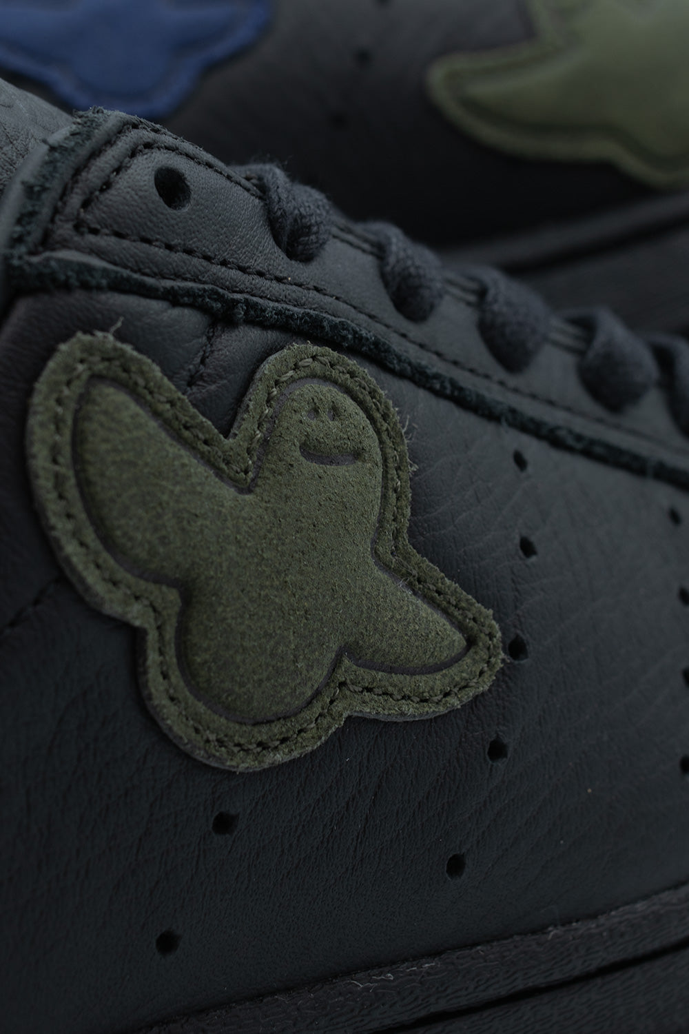 Detailed macro studio view of an olive green suede Shmoo ghost patch edge-stitched onto the black full-grain leather upper of the Adidas Superstar ADV, showing neat topstitching and black waxed cotton laces.