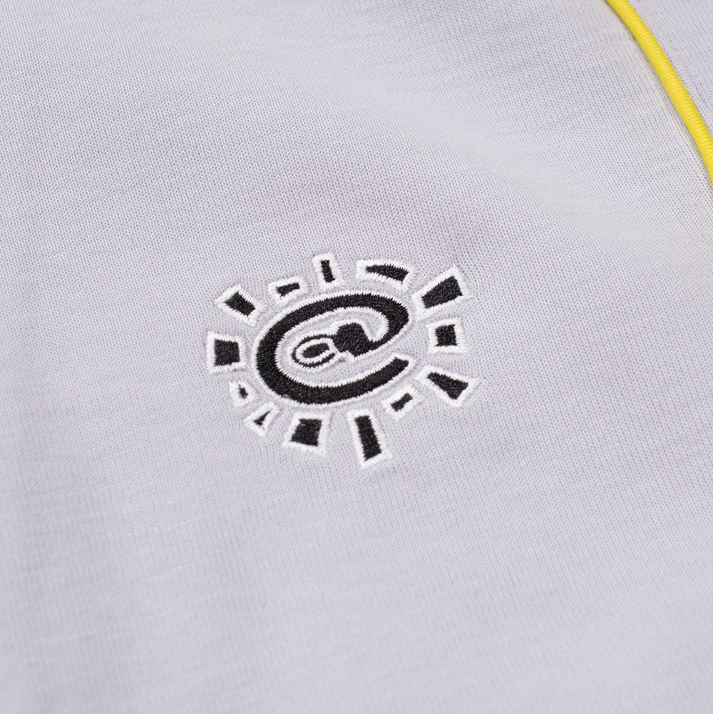 Detailed macro perspective of the chest embroidery on the adidas x ADWYSD Full Zip Hoodie, showing the black satin-stitch thread forming the signature ADWYSD sun emblem with a centered "@" symbol outlined in white thread, set against smooth grey cotton-poly knit fabric next to vibrant yellow piping.