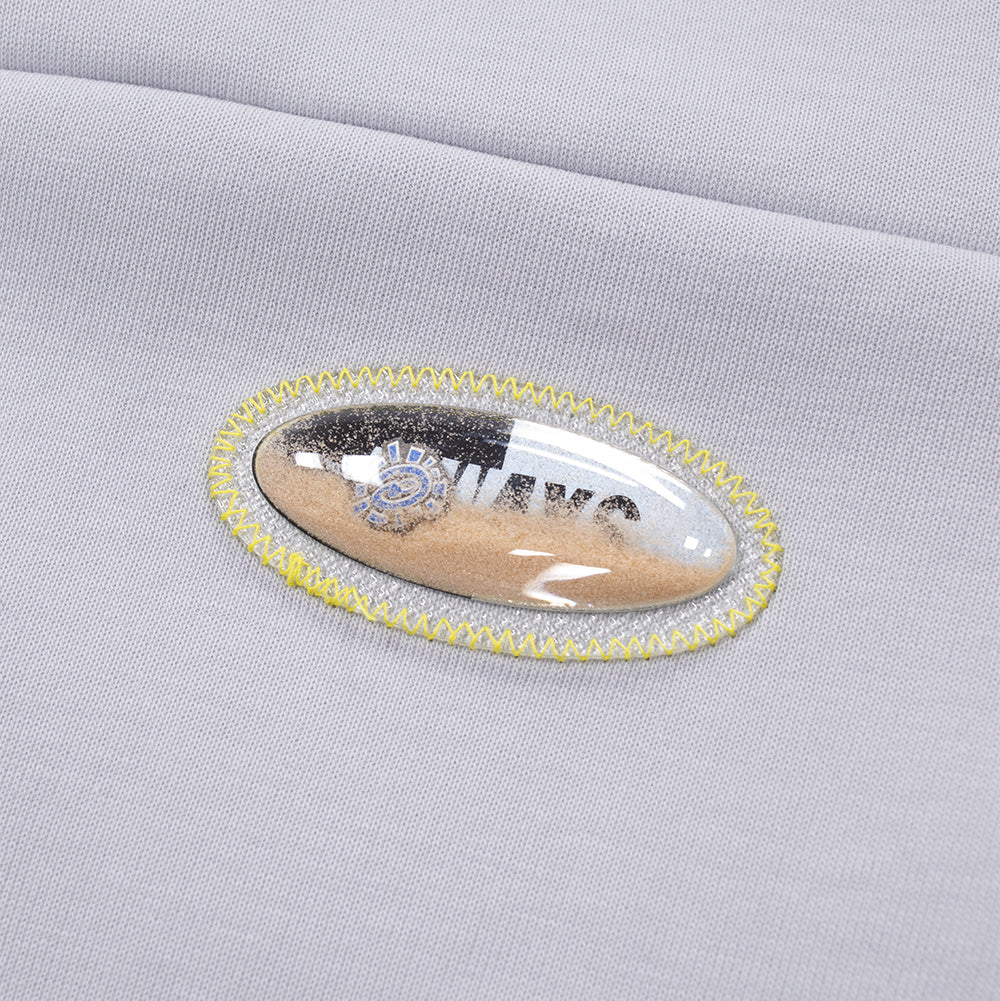 Extreme macro close-up of the novelty badge on the adidas x ADWYSD Full Zip Hoodie, detailing a clear oval bubble patch filled with genuine beach sand and liquid displaying floating branding, secured by thick neon yellow zigzag perimeter stitching on light grey fleece.