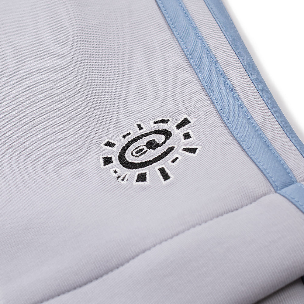 Detailed macro studio close-up of the lower leg embroidery on the adidas x ADWYSD Pant, displaying the dense black satin-stitch thread forming the radiating sun icon with a centered "@" symbol outlined in white thread, set on smooth light grey performance double-knit fabric bordered by the baby blue Three Stripes.