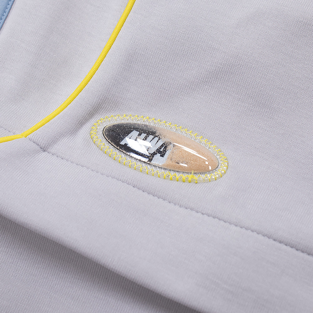 Extreme macro close-up of the novelty badge on the knee flap of the adidas x ADWYSD Pant, featuring a clear oval bubble capsule filled with natural beach sand and liquid displaying floating black text, secured with bright neon yellow zigzag perimeter stitching alongside neon piping.