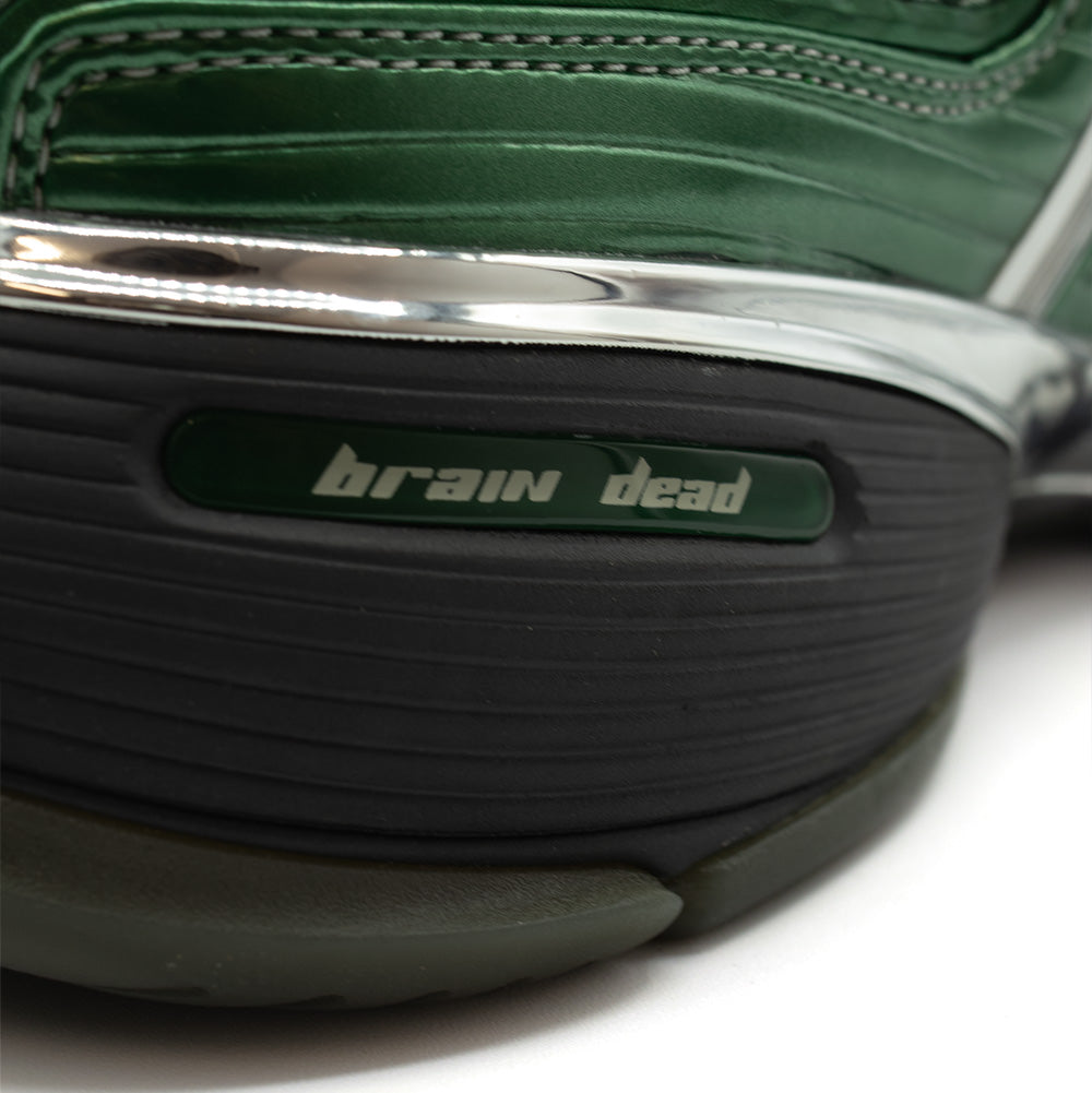 Extreme macro close-up of the lateral rear heel on the adidas x Brain Dead Barricade V sneaker, highlighting the horizontally grooved carbon black midsole section featuring an inset dark green oval badge stamped with silver "brain dead" lowercase typography, bordered above by the mirror-finish metallic chrome heel trim and ribbed emerald green synthetic upper with tonal stitching.