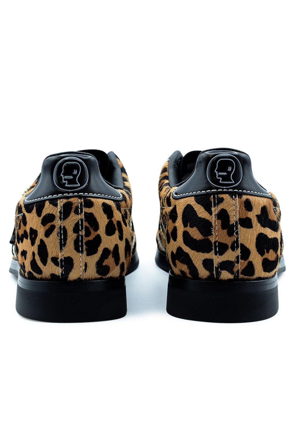 Rear studio shot of the heels of the Adidas X Brain Dead Bowling sneakers, highlighting the textured animal print pony hair heel counters, smooth black leather heel mustache tabs debossed with the Brain Dead head outline logo, white contrast stitching, and raised black rubber block heels.