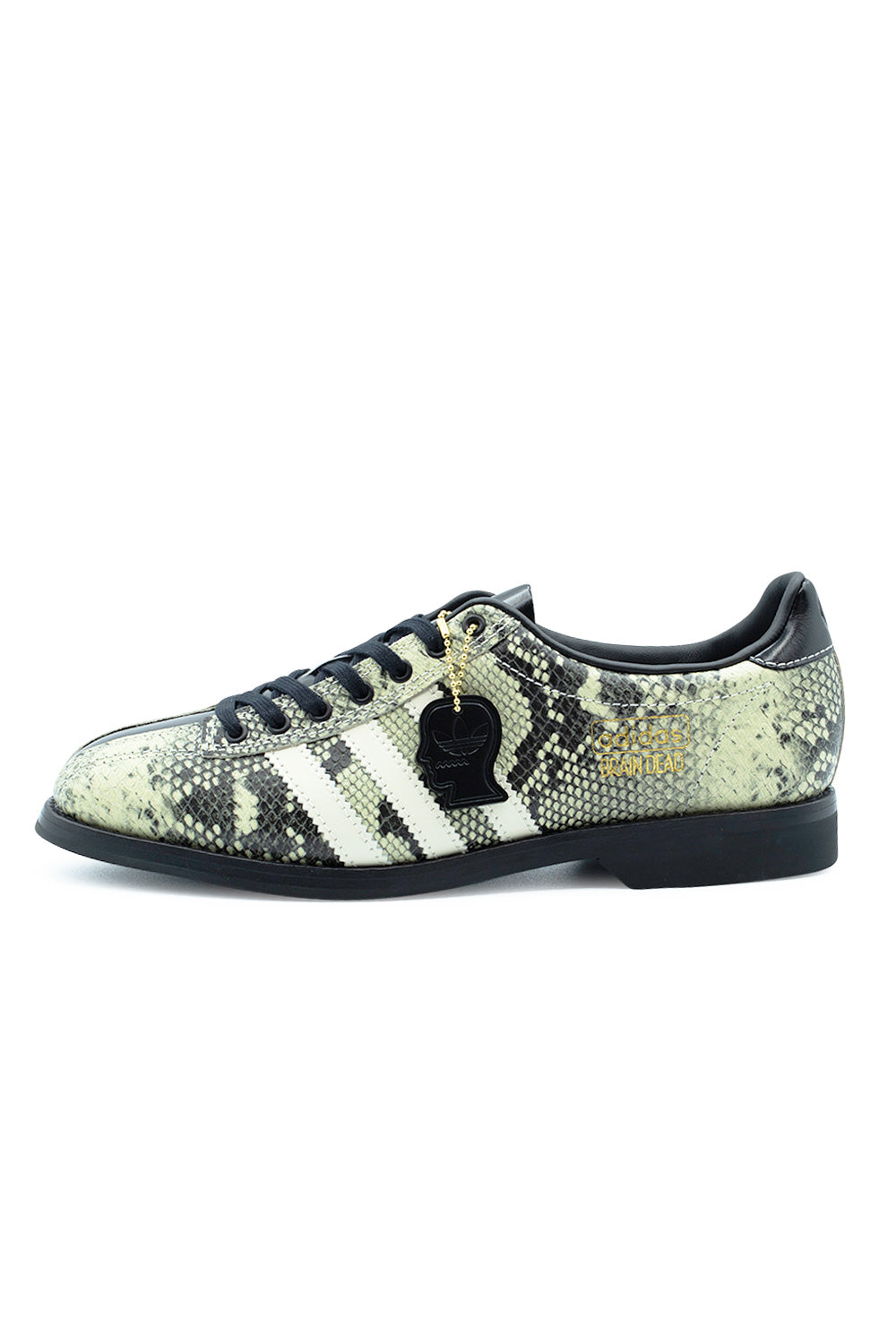 Lateral profile view of the Adidas X Brain Dead Bowling shoe, showcasing the printed snakeskin textured leather upper in light cream and black, off-white serrated Three Stripes, flat black laces, gold-foil Adidas Brain Dead branding, a dangling black metal Brain Dead head hangtag, and a flat black rubber outsole with a distinct raised bowling heel.