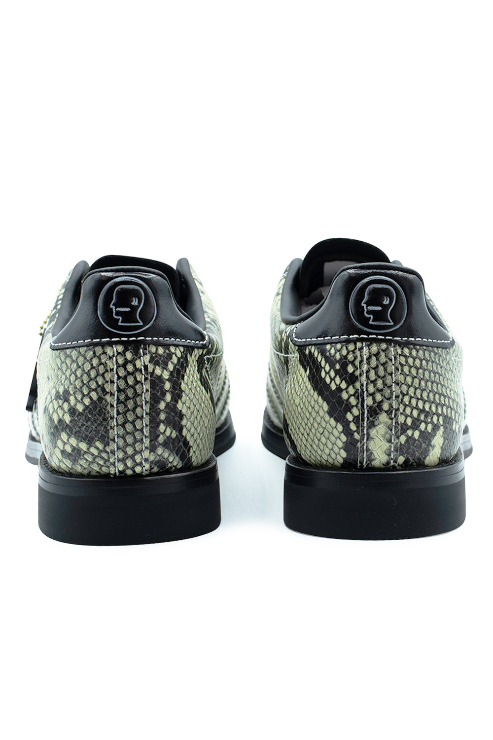 Rear perspective shot of the heels of the Adidas X Brain Dead Bowling sneakers, highlighting the textured snakeskin leather quarters, smooth black leather heel tabs debossed with the white Brain Dead head outline logo, white welt stitching, and solid black raised rubber heels.