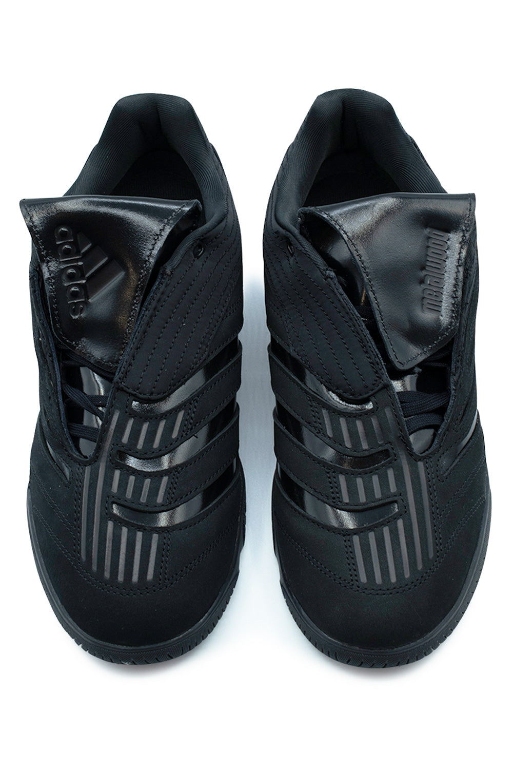 Top-down view of a pair of Adidas X Metalwood Studio Predator Sala shoes in Core Black, showcasing the textured Predator forefoot strike zone ribs, asymmetrical lacing covered by large glossy fold-over tongues embossed with the Adidas logo on the right shoe and Metalwood branding on the left.