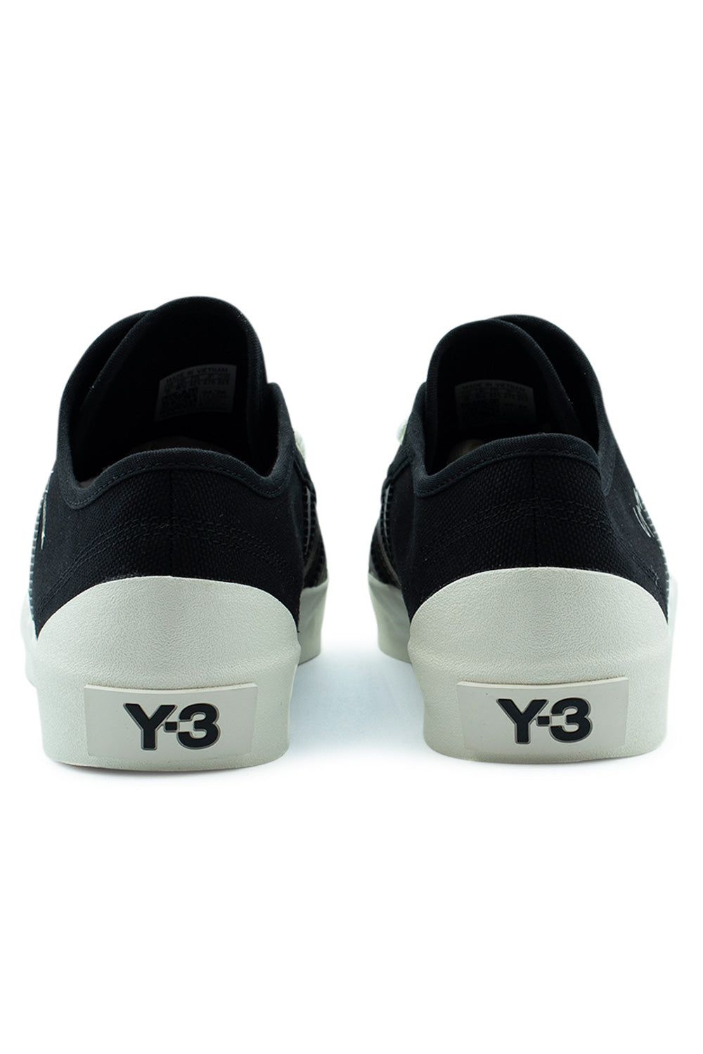 Rear view of the heels of the Adidas Y-3 Nisi Lo shoes in Black and Off White, displaying the black canvas heel counter with fine tonal stitching, elevated off-white rubber midsole wrapper, and a bold black embossed Y-3 logo block on the heel patch.