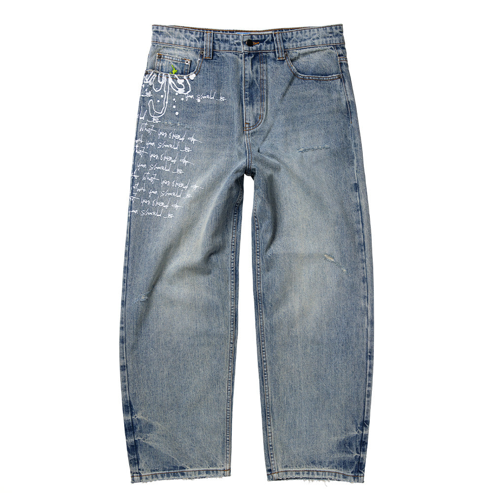 Front flat lay view of the Always Do What You Should Do Waza Jeans in washed light blue denim, showcasing a relaxed fit with slightly tapered legs, classic five-pocket styling, subtle distressed abrasions on the left thigh, custom metal waistband hardware, and prominent white graffiti-style bubble lettering and repetitive script graphics running down the right leg.