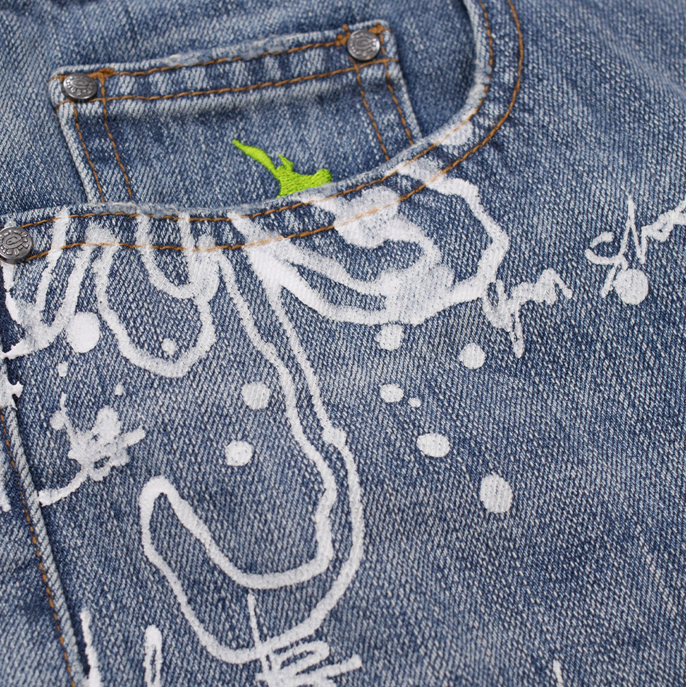 Detailed macro close-up of the front right pocket on the Always Waza Jeans, displaying the bold white paint-splatter graffiti script on washed denim, custom metallic circular rivets, and a lime-green embroidered silhouette of New Zealand peeking out from the internal coin pocket.
